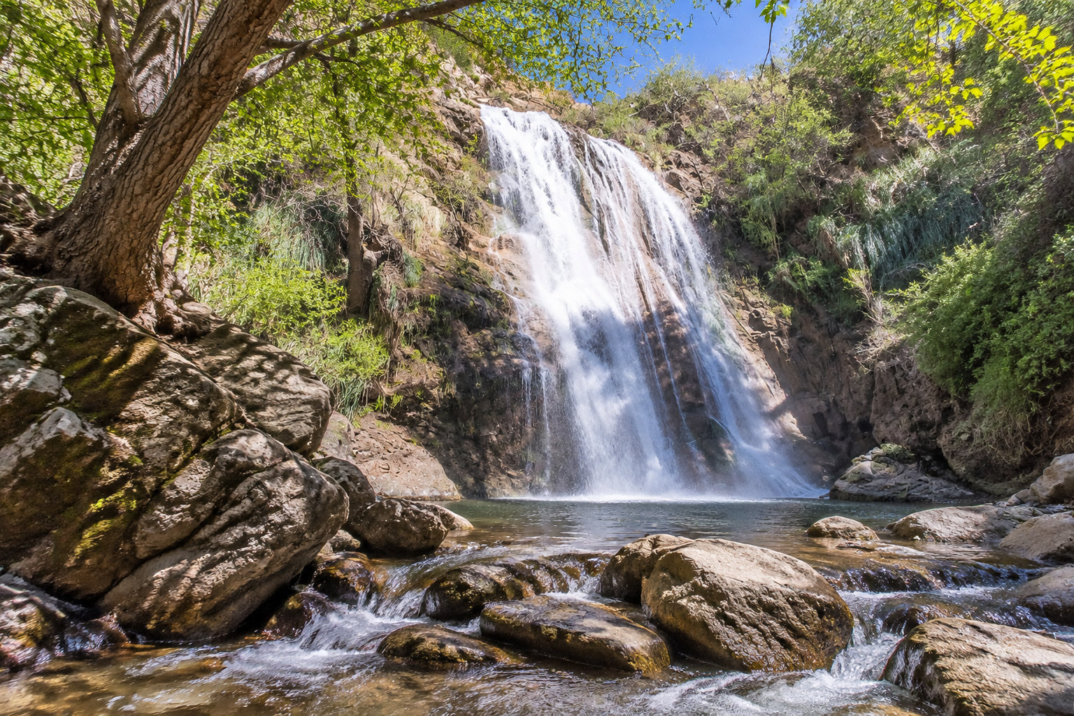 A waterfall cascades over a rocky cliff into a pool surrounded by trees and boulders.