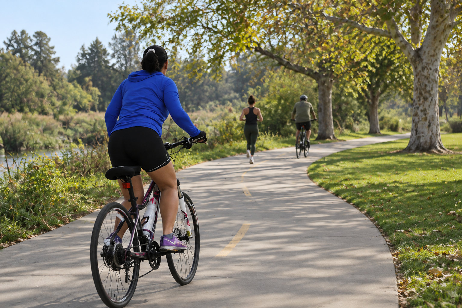 A woman rides a bicycle along a shaded park path, with a jogger and another cyclist ahead.