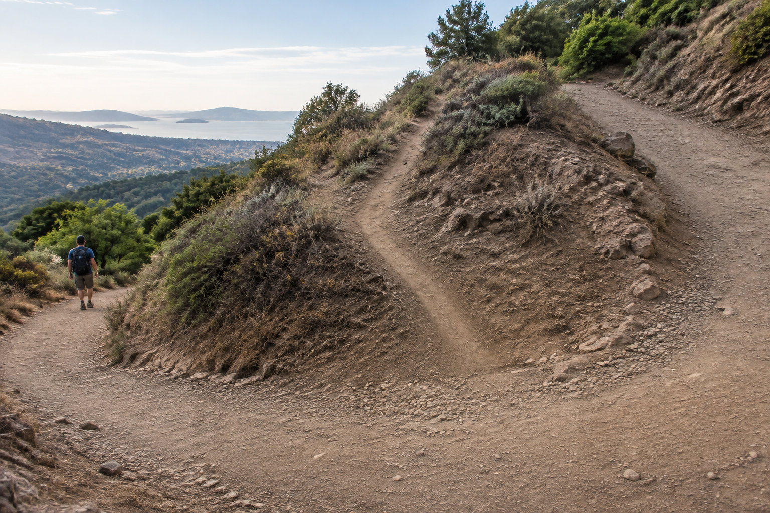 A hiker walks along a winding dirt trail on a rocky hillside overlooking a body of water and distant hills.