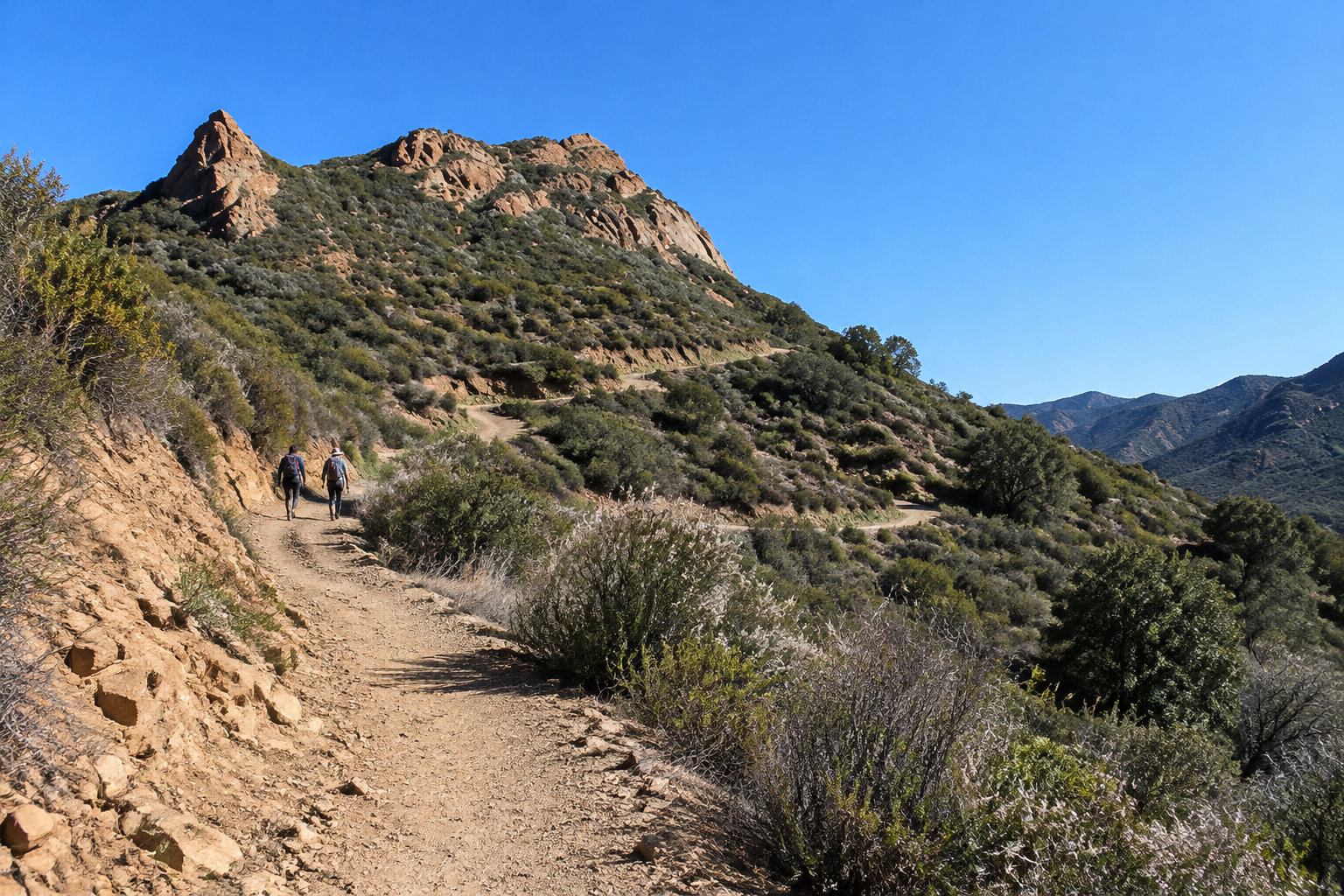 Two hikers walk along a winding dirt trail through scrub-covered hills beneath a clear blue sky.